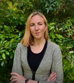 Photo of blond woman in a grey sweater standing with arms crossed in front of green foliage.