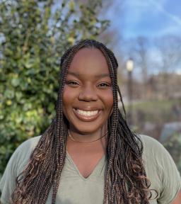 Headhsot smiling in light olive green t-shirt with gold choker chain in front of blurred park background