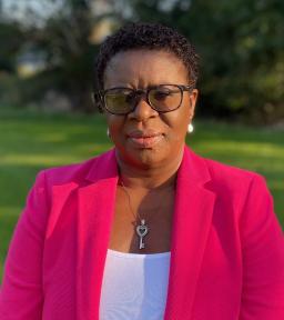 Headshot with glasses and a pink jacket over a white shirt in front of greenery