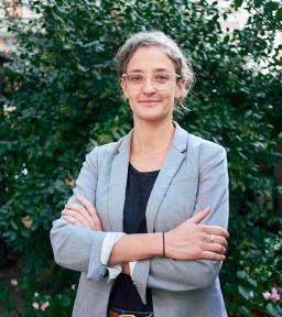 Head shot with arms crossed, wearing a light gray blazer over a black shirt, standing in front of green foliage.