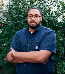 Headshot, arms crossed, wearing navy blue short-sleeved button-down shirt, standing in front of green foliage.