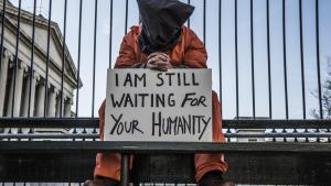 Protester in detainee garb holding sign, "I am still waiting for your humanity"