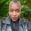 Headshot of Sunyata with silver hoop earrings and a black motorcycle jacket in front of a mottled green background